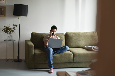Man using laptop and calling while sitting on sofaの写真素材