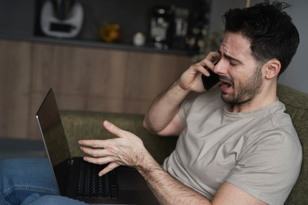 Frustrated man using laptop and calling while sitting on sofaの写真素材