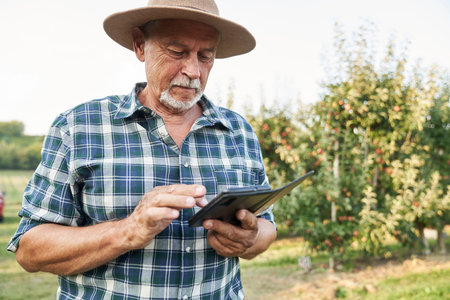 Senior farmer browsing mobile phone while standing on apple farmの写真素材