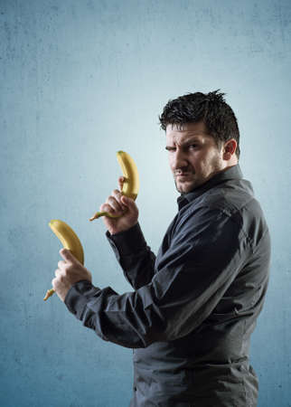 Angry man holding bananas like pistols. He looking at camera with rage face expression.