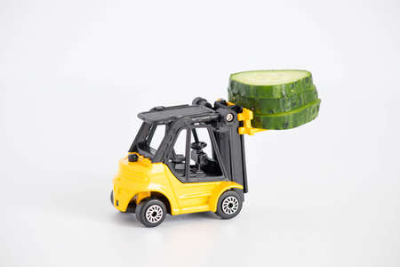 A toy loader or construction crane in bright yellow carries a piece of cucumber and a piece of black...
