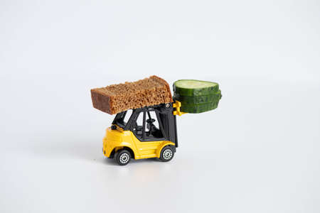 A toy loader or construction crane in bright yellow carries a piece of cucumber and a piece of black freshly baked bread. Close-up miniatures on a white background.の写真素材