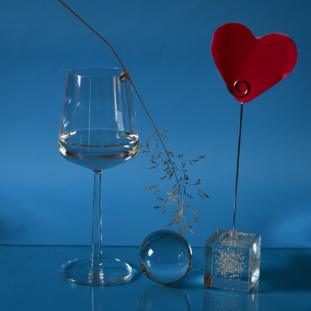 Red heart, glass with a little water, dried straw and glass ball on shiny surface against blue background.の写真素材