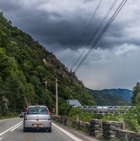 Minivan passing on a mountain road with stone railing on the right side, forest covered mountain and electricity pole in the background with dark sky aboveの写真素材