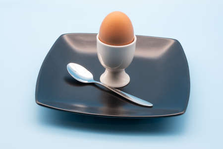 egg in a white cup with a spoon on a black plate isolated against a ight blue backgroundの写真素材
