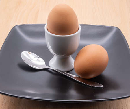 two eggs a spoon and an egg cup on a black plate isolated on a brown chopping boardの写真素材