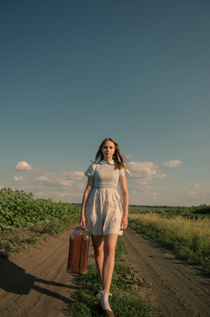 Cute girl goes on a dirt road in field with a suitcase in handの写真素材