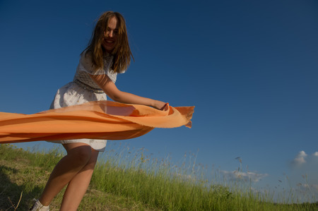 BEAUTIFUL BLONDE GIRL LAUGHS AND DANCES OUTDOORS WITH AN ORANGE SCARF IN HER HANDS ON A MEADOW DURING SUNSETの写真素材