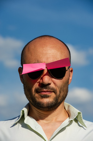 conceptual portrait of a businessman in modern sunglasses pasted with pink stickers against the blue sky. The wind opens and closes the stickers on the glasses.の写真素材