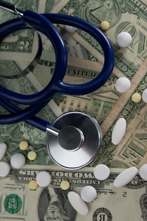medical stethoscope lies against a background of dollar bills and pills.