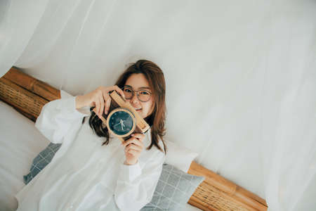 Portrait of happy Asian beautiful woman Smiling in white shirt posing on bed. Taking pictures from a clock that looks like a photo camera. People lifestyle concept. copy space. looking at camera.の写真素材