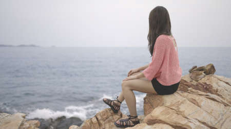 A beautiful woman in pink sweater sits on a rock above the sea, looking at the ocean. Girl traveler, backdrop of the sea and sky. thinking, freedom, solitude, relaxed, goal concept. copy space.の写真素材