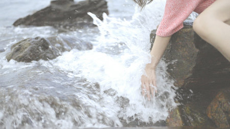 Girl sitting on the rocks. Young woman's hand touching the water in the sea, catching the wave with her hand. Splashing Waves at Blowing Rocks.の写真素材