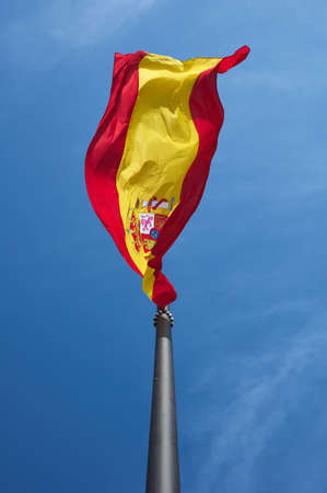 detail of the Spanish flag flying on a large black mast against a blue sky.