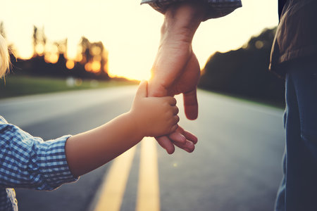 Close-up of a child's and adult's hands holding each other against a blurred sunset backdrop, capturing warmth, intimacy, and connection in a gentle, protective grip.の素材