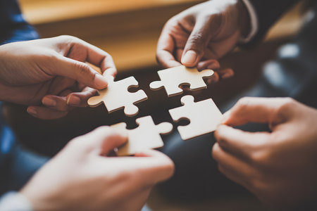 Close-up of hands connecting white jigsaw puzzle pieces against a blurred brown background. Warm natural lighting enhances the soft bokeh effect.の素材