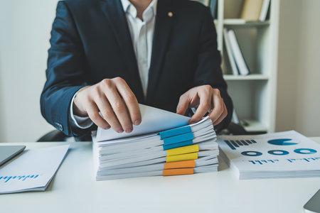 Close-up of hands in a navy suit sorting white documents with colorful tabs on a minimalist office desk, featuring financial charts and a bright, professional ambiance.の素材