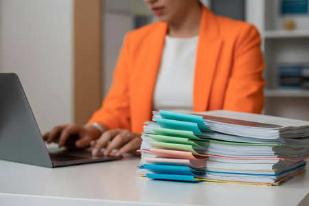 Close-up of a bright orange blazer-clad professional working at a modern laptop, with organized business documents and colorful tabs in a stylish office setting.の素材
