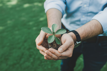 Close-up of hands in a light blue sleeve cradling a small green sprout in dark soil, set against a blurred vibrant green grass background. Soft natural lighting.の素材