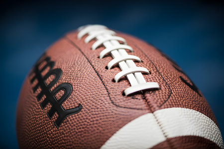 Close-up macro shot of an American football set against a dark blue background, highlighting textured leather, white laces, and dramatic lighting for a striking visual effect.の素材
