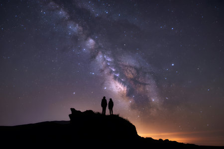 Silhouetted figures on a rocky hilltop under the breathtaking Milky Way, showcasing a vibrant mix of deep blues, purples, and warm orange hues in a dramatic night sky.の素材