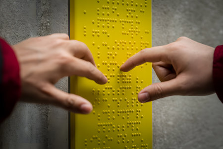 Close-up of two hands in red sleeves gently touching a bright yellow tactile panel with clear braille on a textured concrete background. Emphasizing tactile interaction.の素材