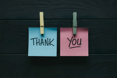 Flat lay of colorful sticky notes on dark wood: 'Thank' in soft blue and 'you' in dusty rose, secured with muted clothespins, showcasing a modern minimalist style. Thank you day.の素材