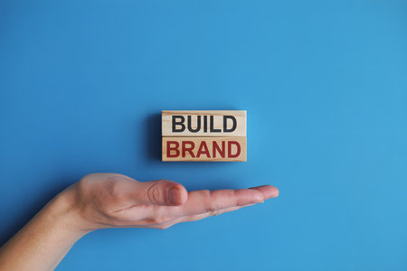 Minimalist flat lay of wooden blocks with BUILD and BRAND on bright blue background, featuring a hand presenting the blocks. Soft lighting enhances warm wood texture.の素材