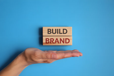 Minimalist flat lay of wooden blocks with BUILD and BRAND on bright blue background, featuring a hand presenting the blocks. Soft lighting enhances warm wood texture.の素材