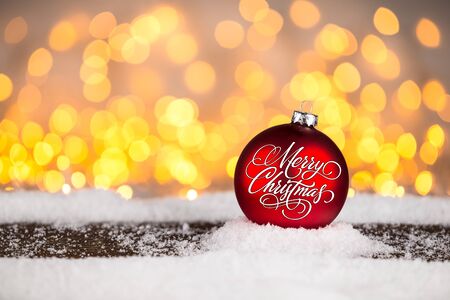 red merry christmas xmas bauble ball on snowy wood plank in front of many bright illuminated lights bokeh backgroundの写真素材