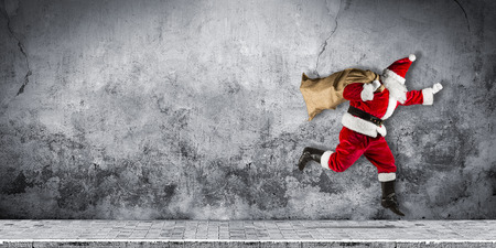 late Santa claus in a hurry with traditional red white costume and bag full of presents running jumping abstract funny christmas xmas concrete background with copy spaceの写真素材