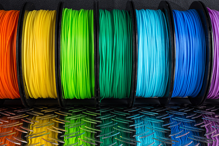 colorful bright row of spool 3d printer pla abs filament plastic material on dark black metal steel...