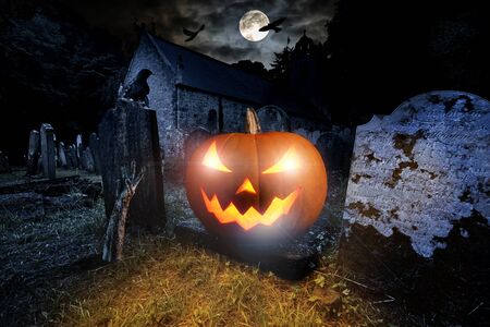 glowing orange halloween pumpkin with scyry zombie hand on old graveyard in front of full moon black raven church dark night spooky horror backgroundの写真素材