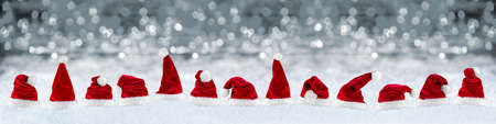 wide panorama row of red white plush santa claus christmas xmas hat on snow in front of silver gray bokeh lights backgroundの写真素材