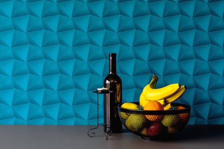 A background with blue wallpaper - colorful space. Black fruit basket in an elegant arrangement. Black candle holder with a candle warmer, black wine bottle as a decoration. Fresh fruits such as banana, orange, pear, and apples.の写真素材
