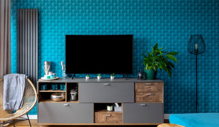 A furnished living room in a modern style. Blue interior of a well-lit room. A dresser with drawers and a large flat-screen TV. Plenty of decorations: a radiator, curtains, a candle lamp, candlesticks, and a green plant in a pot.の写真素材