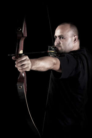 Archer in black on black background aiming with bow and arrow, with focus on bow. 