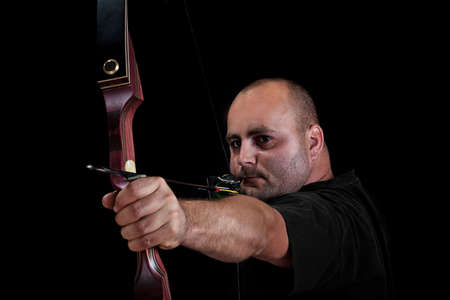 Archer in black on black background aiming with bow and arrow, with focus on bow. の写真素材