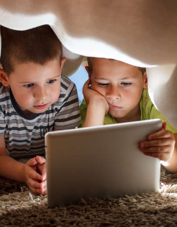 Two children hiding under blanket and holding tabletの写真素材