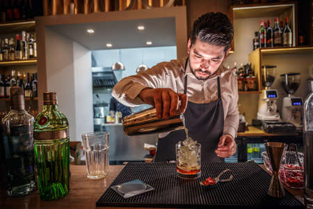 Male bartender is making cocktail pouring alcohol from shaker to glass at bar backgroundの写真素材
