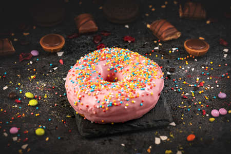 Still life of pink donut decorated with colorful sprinkles on black slate background