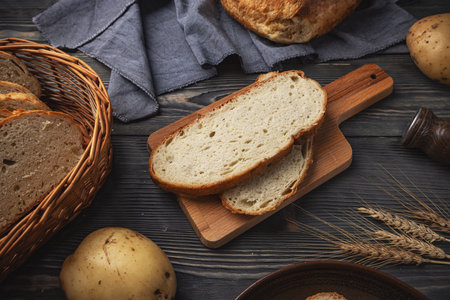 Freshly sliced rustic bread on a cutting board, with potatoes and wheat, on a dark wood surfaceの写真素材