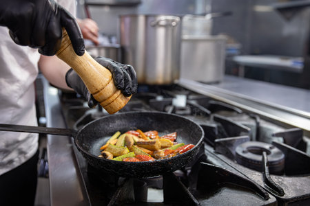 Chef adding spices to grilled vegetables in frying pan on gas stoveの写真素材