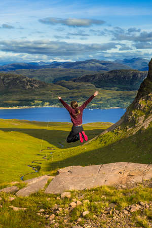 Young Pretty Girl Jumps Happily From Rock In Front Of Spectacular Landscape Of Applecross Pass In...