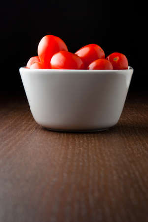 A white bowl of fresh red cherry tomatoes on a wood table against a black background.の写真素材