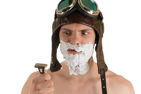 Portrait of an adult man with shaving foam on his face in flight helmet and flying goggles looking at razor in his handの写真素材