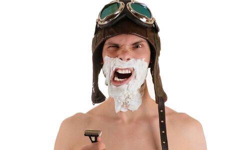 Portrait of screaming man with narrowed eyes with shaving foam on his face in flight helmet and flying goggles with razor in his handの写真素材