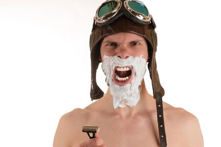 Portrait of screaming man with shaving foam on his face in flight helmet and flying goggles with razor in his handの写真素材