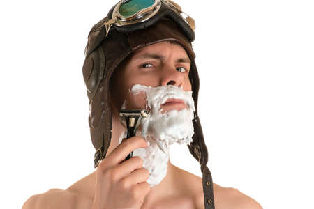 portrait of a man shaving with shaving foam on his face in a flight helmet and flying goggles arrogantly looking at the cameraの写真素材