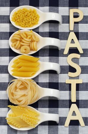 Pasta written with wooden letters on checkered cloth and uncooked pasta on porcelain spoonsの写真素材